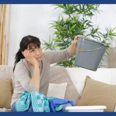 Woman sitting on couch holding bucket to catch water from ceiling leak, looking frustrated with roof damage