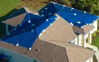 Aerial view of residential Florida home with blue emergency tarps covering damaged roof sections after hurricane, showing multiple roof slopes requiring storm damage repair assessment for 25% rule calculation
