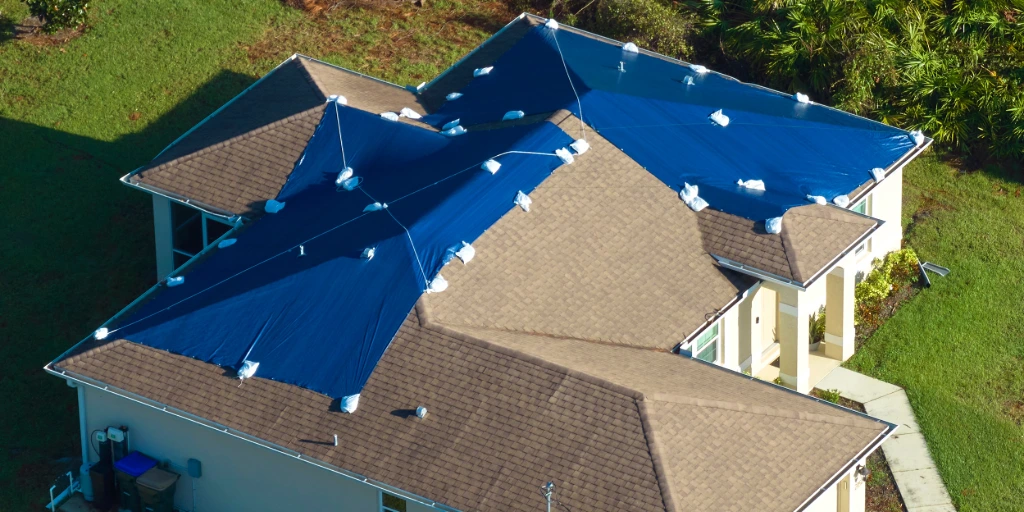 Aerial view of residential Florida home with blue emergency tarps covering damaged roof sections after hurricane, showing multiple roof slopes requiring storm damage repair assessment for 25% rule calculation